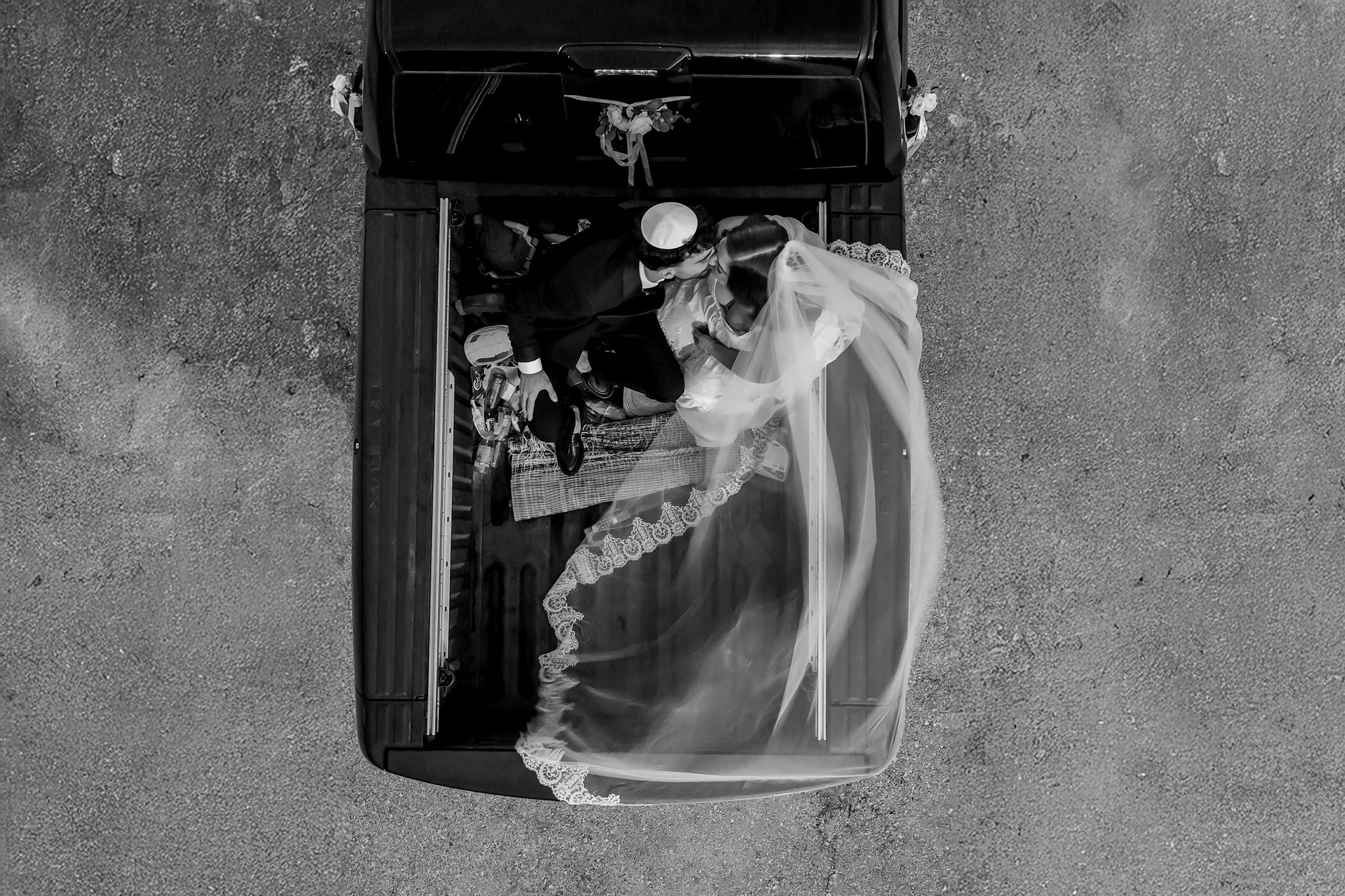 A black and white overhead photograph capturing a newly married couple sharing an intimate moment in the bed of a classic black pickup truck. The groom, wearing a dark suit and a white kippah, is leaning in towards the bride, who is in a white wedding dress with a long, flowing, lace-trimmed veil. They appear to be kissing or embracing tenderly. The truck bed contains items like woven baskets, suggesting a journey or celebration, and the vehicle is subtly adorned with floral arrangements at the back. The truck is parked on a textured asphalt or concrete surface, making the couple the central focus of this unique and romantic scene.