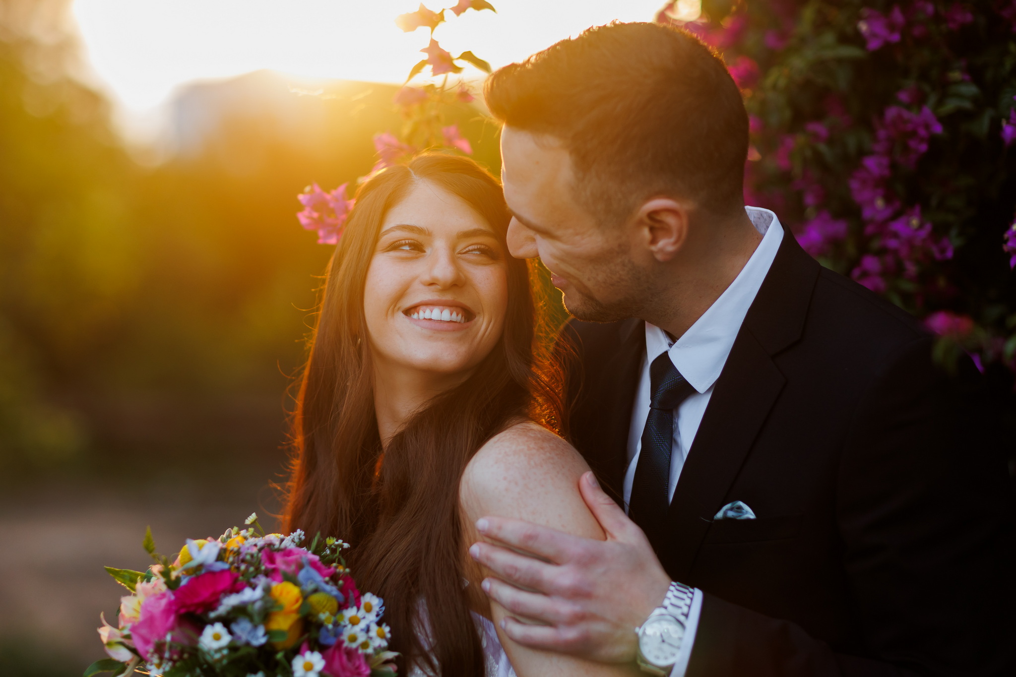 A joyful couple embracing outdoors during golden hour. The woman, with long dark hair and a radiant smile, looks up lovingly at the man. She holds a vibrant bouquet of pink, yellow, blue, and white flowers. The man, wearing a black suit with a dark tie and a silver watch, gazes down at her with a gentle smile, his arm around her shoulder. The background is softly blurred with warm, golden sunlight on the left and lush purple bougainvillea flowers on the right, creating a romantic and intimate atmosphere.