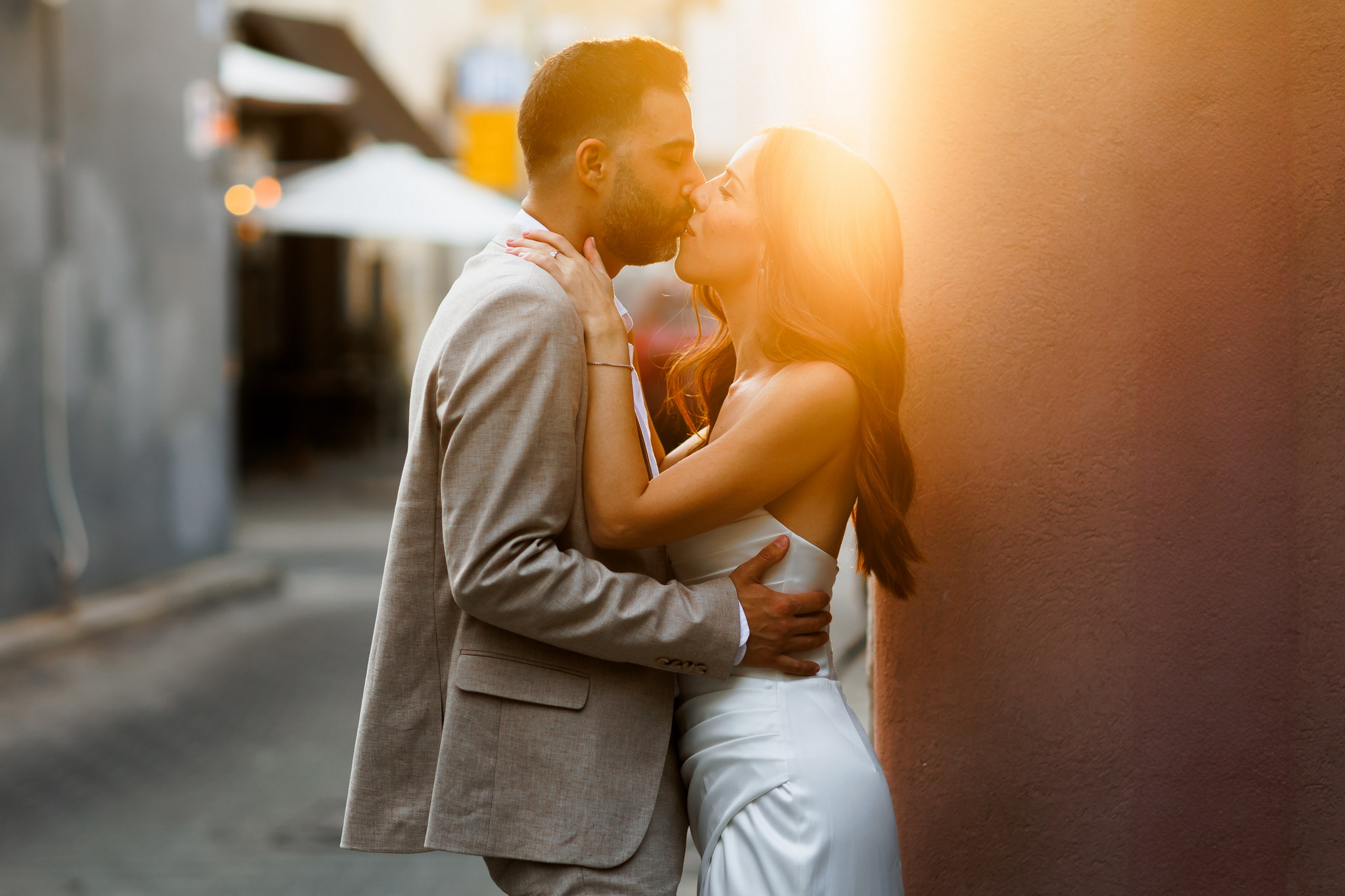 A romantic couple, dressed for a wedding, embraces and kisses passionately in a sunlit urban alley. The man wears a light beige suit and has a short beard, while the woman has long, wavy brown hair illuminated by a strong golden light, and wears a white strapless dress. Her hand with a ring is visible on the man's neck. A bright golden lens flare from the upper right bathes the scene in warm light, creating an intimate atmosphere. The background is softly blurred, showing hints of city architecture on the left and a textured warm-toned wall on the right.