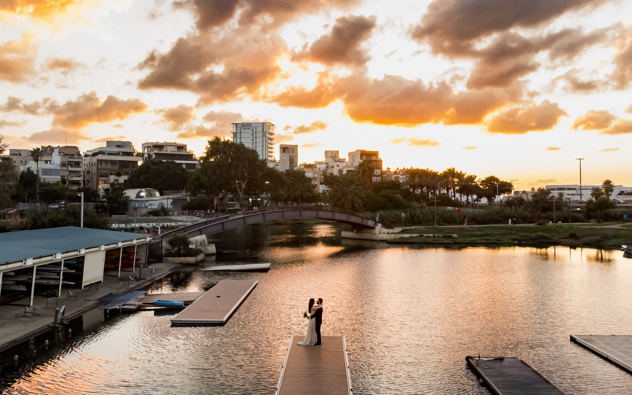 An aerial view of a romantic couple in wedding attire embracing on a long, wooden dock that extends into tranquil water. The woman, in a light wedding gown and holding a bouquet, faces the man, who is in a dark suit. The calm water reflects the vibrant orange and golden hues of a dramatic sunset sky, which is filled with dark and bright clouds. In the background, a pedestrian bridge arches over the water, leading to a cityscape featuring various buildings, lush trees, and palm trees lining the distant bank. The entire scene is bathed in the soft, warm glow of the setting sun, creating a serene and picturesque atmosphere.