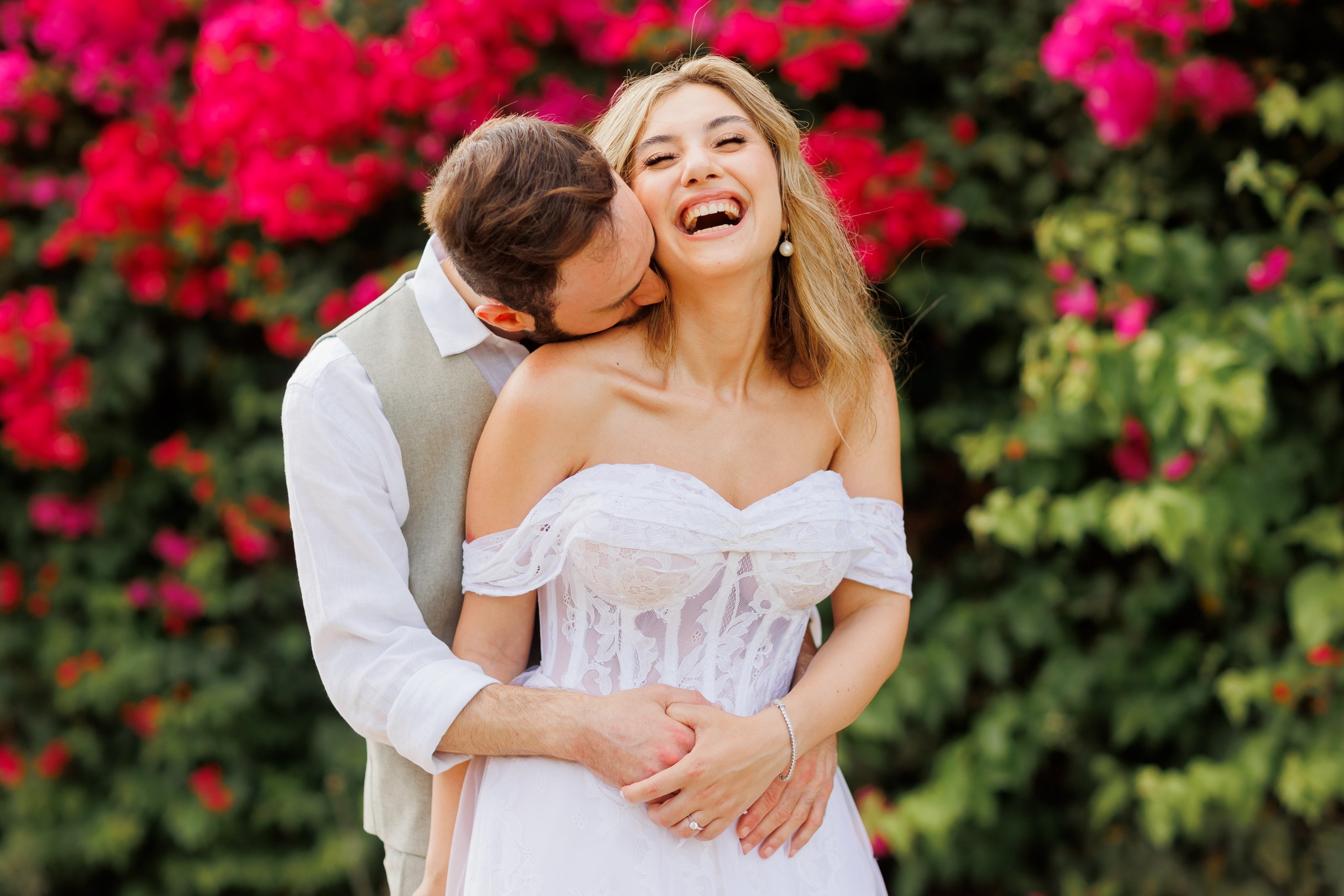 A joyful couple in wedding attire embracing against a vibrant backdrop of lush green foliage and bright pink bougainvillea flowers. The bride, with long blonde hair and an off-the-shoulder white lace gown, smiles broadly with her head tilted back, revealing an engagement ring and pearl earrings. The groom, in a white shirt and light vest, gently kisses her neck, with his arms wrapped around her waist, creating an intimate and romantic scene.