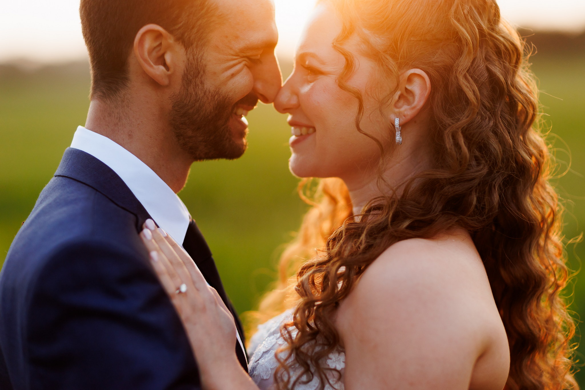 A close-up shot of a joyous wedding couple embracing during golden hour. The groom, with a beard and dark suit, is on the left, smiling broadly with his eyes closed as his nose gently touches the bride's. The bride, on the right, has long wavy reddish-brown hair, glowing with the warm backlight, and wears a light-colored dress with a sparkling diamond earring. Her left hand, adorned with an engagement ring, rests gently on the groom's shoulder as she smiles with her eyes closed. The warm, golden sunlight bathes their faces and hair, while a soft, blurred green field provides a natural, romantic background.