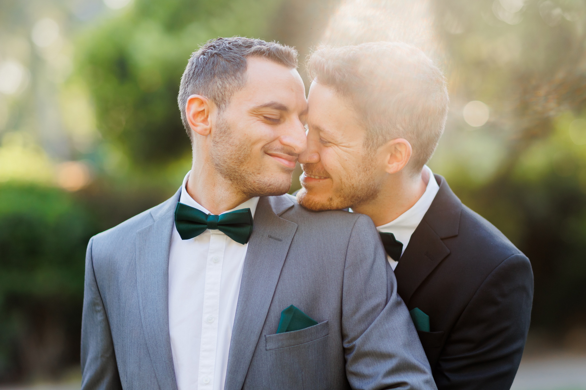 A joyful same-sex couple embracing tenderly outdoors. The man on the left wears a grey suit with a dark green bow tie and matching pocket square, his forehead gently touching his partner's. The man on the right, in a black suit with a dark green pocket square, has his head nestled against the other's shoulder. Both have their eyes closed and are smiling softly, conveying deep affection. The background is a softly blurred expanse of vibrant green foliage, illuminated by a warm, natural light creating a gentle sun flare on the right side.
