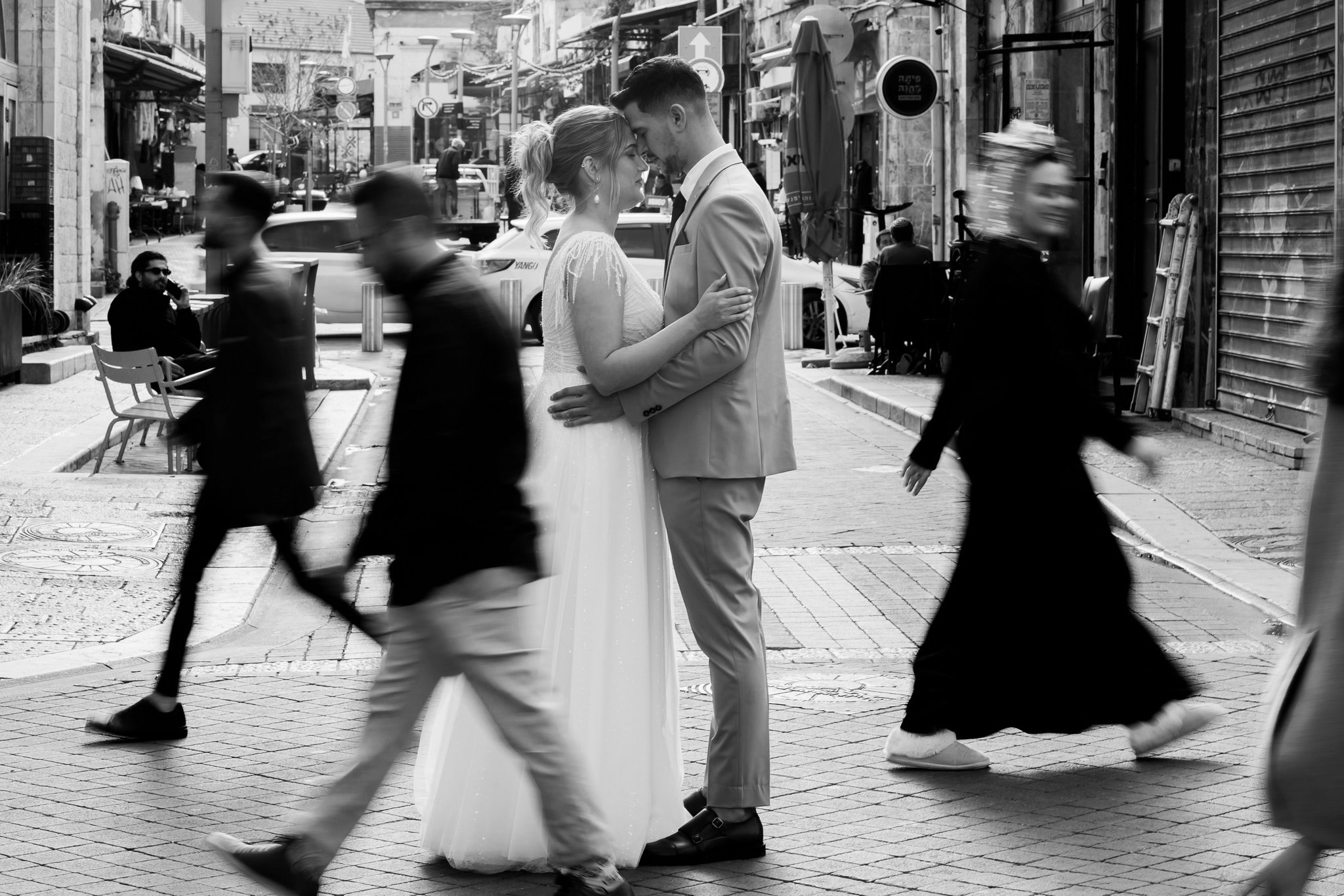 A black and white street photograph capturing an intimate moment between a newlywed couple embracing in the bustling center of an urban street. The bride, in a sparkling white gown with a fitted bodice and full skirt, and the groom, in a light-colored suit and dark tie, stand forehead-to-forehead with their eyes closed, locked in a gentle hug. They are perfectly still, creating a serene focal point amidst the blurred motion of several passersby on the cobblestone pavement, suggesting a fast-paced city environment. In the background, there are street signs, parked cars, and old buildings housing storefronts and outdoor cafes, with a few people visible sitting or standing. The high contrast and monochrome tones emphasize the timelessness and emotional depth of the couple's connection despite the surrounding activity.