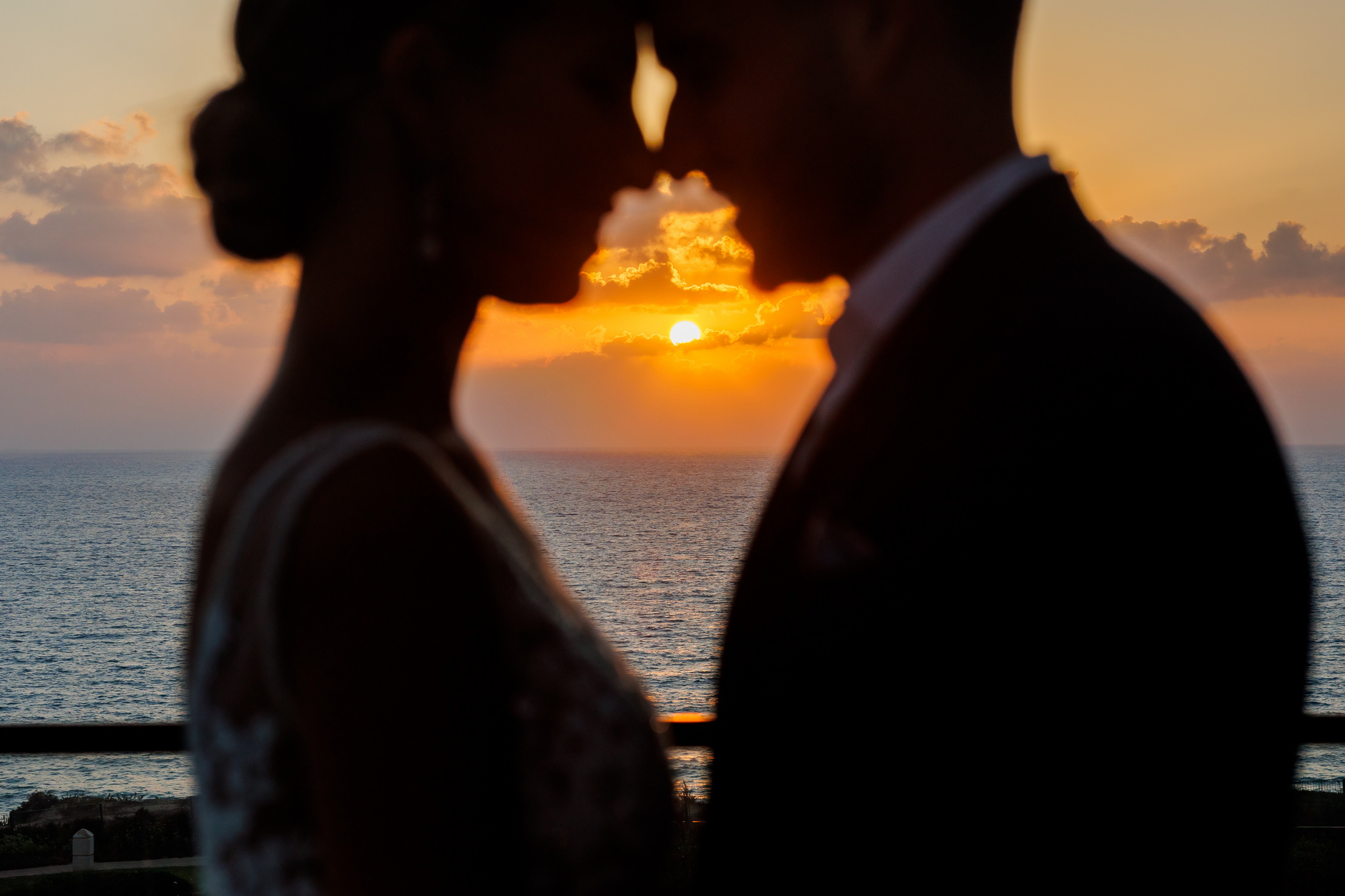 A romantic image capturing a silhouetted couple intimately close, with their foreheads touching, against a stunning sunset over the ocean. The woman, possibly a bride in a wedding dress, and the man, likely a groom in a suit, are framed by the vibrant, fiery orange and yellow sun, surrounded by soft clouds. The calm, dark blue sea stretches into the distance beneath the dramatic sky, creating a warm, peaceful, and deeply romantic atmosphere from an elevated viewpoint.