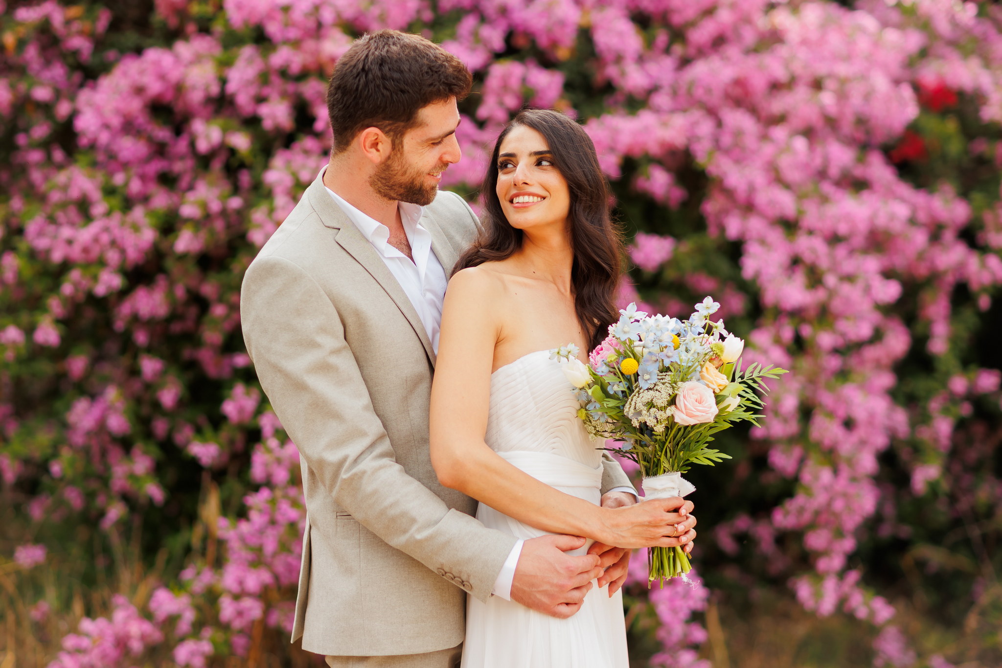 A happy couple in elegant attire embracing against a backdrop of vibrant pink and purple bougainvillea flowers. The man, with a beard and wearing a tan suit and white shirt, gently holds the woman from behind, gazing lovingly at her. The woman, wearing a white strapless dress with long dark hair, holds a colorful bouquet of light blue, pink, yellow, and white flowers, smiling joyfully and looking back at the man. The background is softly blurred, highlighting their affectionate interaction.