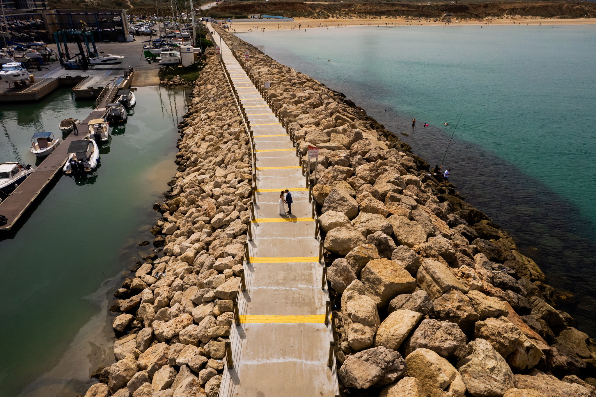 An aerial view captures a newlywed couple in wedding attire standing near the bottom-center on a long, concrete and rock breakwater, stretching into the sea under a bright, clear sky. The bride wears a flowing white dress and the groom a dark suit, facing each other. To the left of the breakwater, a bustling marina is visible with numerous boats docked and a large blue crane. To the right, the water is a vibrant turquoise, leading to a wide sandy beach dotted with people sunbathing and swimming, backed by a distant low brown hill. A few individuals can be seen swimming or fishing from the rocky side of the breakwater.