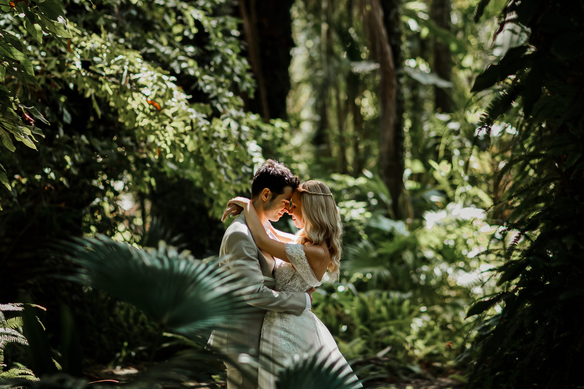A romantic wedding couple embraces intimately in a vibrant green, lush tropical setting. The groom, dressed in a light-colored suit, gently holds the bride who is adorned in an elegant white lace off-the-shoulder gown and a braided hairstyle. Their foreheads touch as soft sunlight filters through the dense foliage, creating a serene and natural backdrop around them.