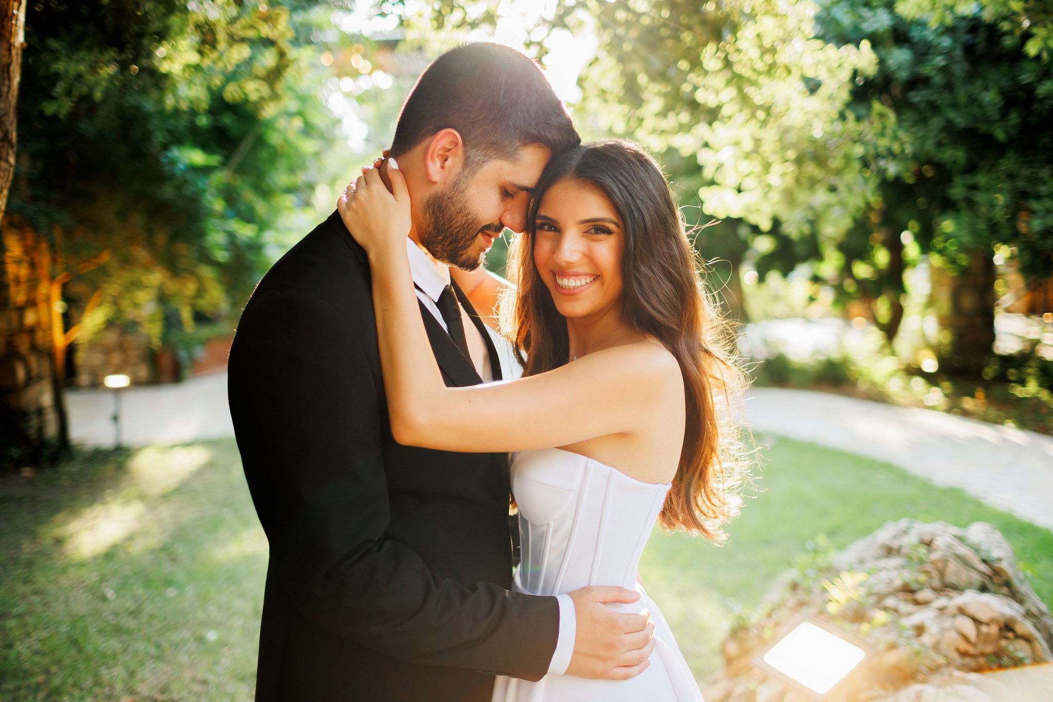 A joyful wedding couple embracing closely in a sunlit outdoor garden. The bride, wearing a white strapless gown, smiles brightly at the camera, while the groom in a black suit gently holds her, resting his head near hers. Warm, soft sunlight streams through the lush green trees and foliage in the background, creating a romantic and natural atmosphere.