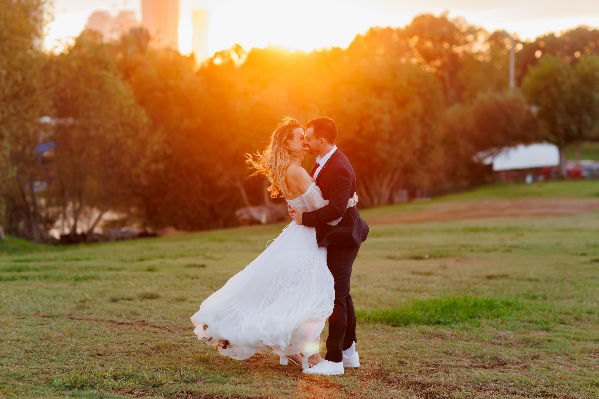 A newlywed couple embraces lovingly in a sun-drenched, grassy field during golden hour. The bride, with windswept blonde hair, wears a flowing white off-the-shoulder wedding gown and white sandals. The groom, in a dark suit and white sneakers, holds her close as they share an intimate moment. The background features warm, glowing golden sunlight filtering through lush green trees, creating a radiant and romantic atmosphere.