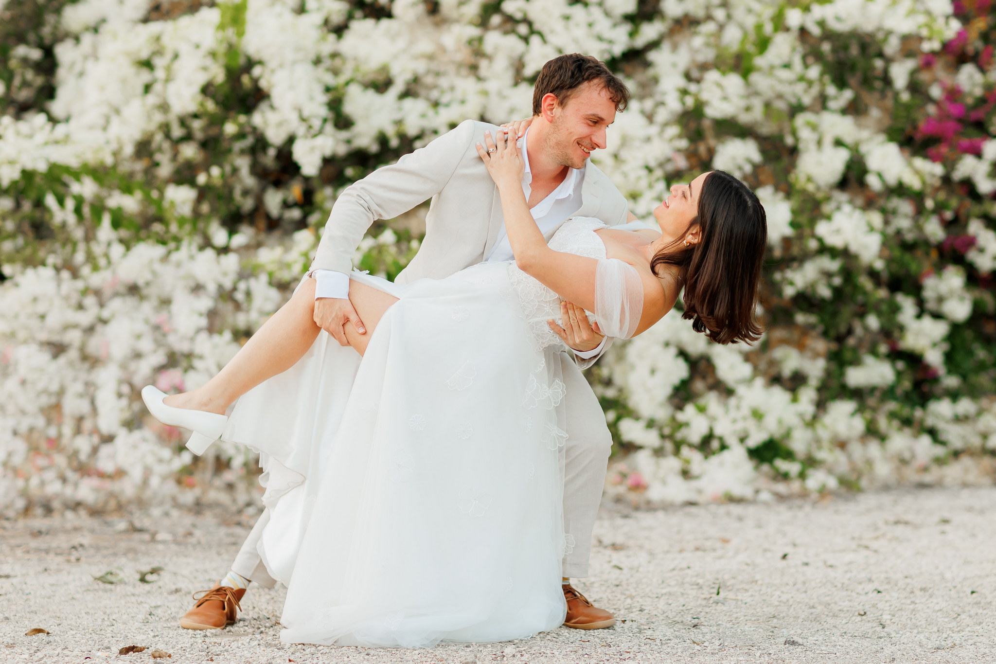 A joyful couple in wedding attire captured outdoors in a romantic dip pose. The groom, wearing a light beige suit and brown shoes, gently holds his bride, who is dressed in a flowing white strapless wedding gown with sheer off-the-shoulder sleeves and white low-heeled shoes. Both are smiling brightly and looking lovingly into each other's eyes. They are standing on light-colored gravel, with a vibrant, soft-focus background of dense white flowering bushes, interspersed with touches of pink or purple blossoms, creating a celebratory and natural atmosphere.