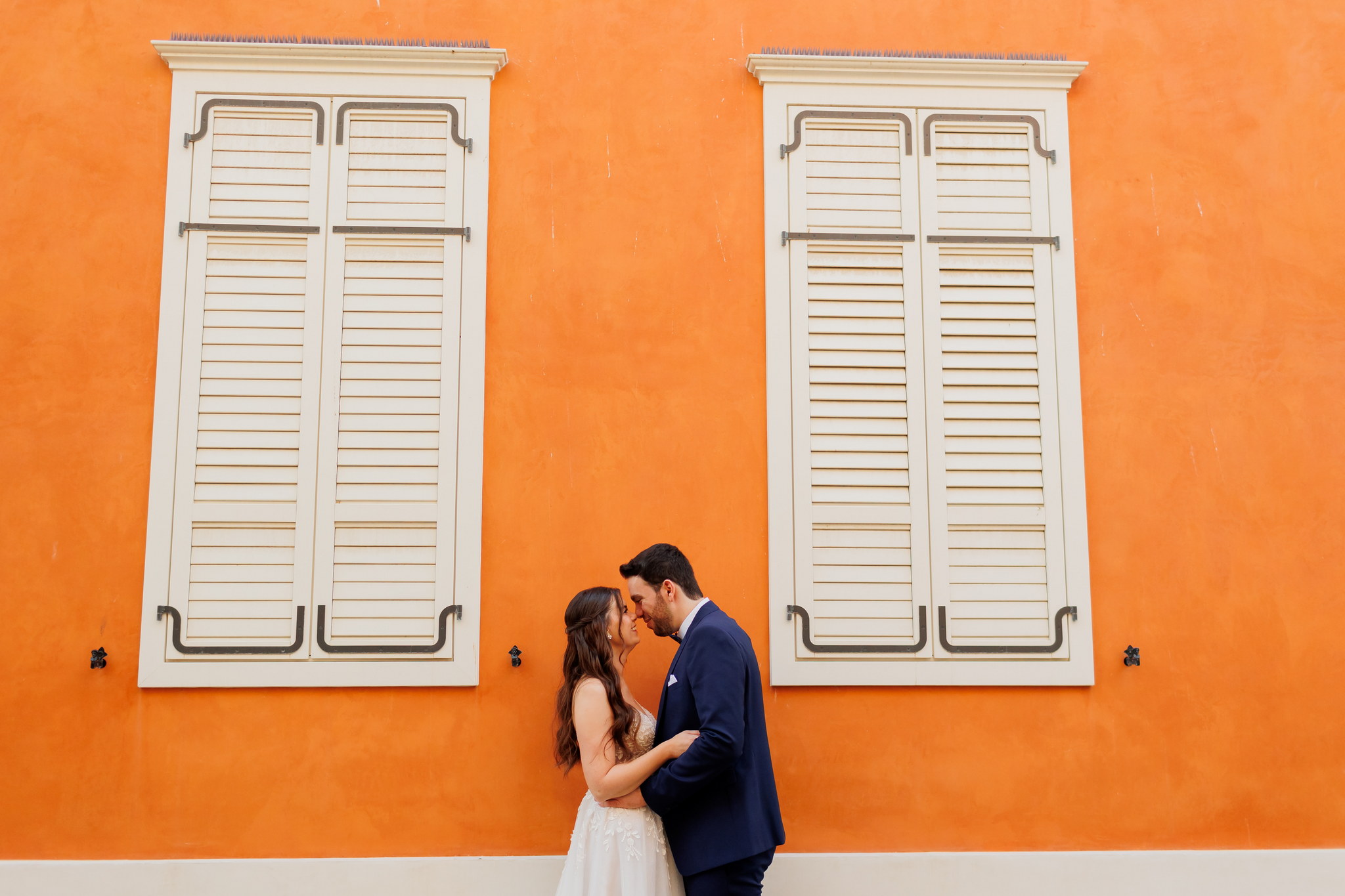 The image shows a joyful wedding couple embracing intimately against a vibrant orange stucco wall. The bride, with long wavy dark hair, is wearing a white embroidered strapless wedding gown, smiling up at the groom. The groom, dressed in a navy blue suit and bow tie, leans in towards her as if for a kiss. They are centered between two large, closed white louvered shutters with decorative dark metal hinges, which stand out against the vivid orange background. A narrow white baseboard runs along the bottom of the wall.