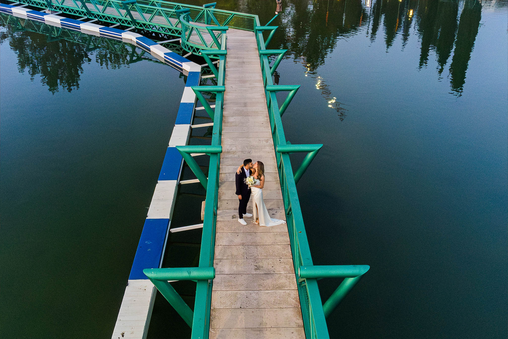 An aerial view captures a newlywed couple sharing a romantic kiss on a modern wooden and teal-green pedestrian bridge extending over dark, calm water. The groom, in a dark suit and white sneakers, gently holds the bride, who is dressed in an elegant white sleeveless gown with a leg slit and holds a bouquet of white and yellow flowers. The calm water reflects hints of distant trees or lights, and a section of blue and white striped floating elements runs alongside the bridge on the left. The scene evokes a serene and intimate atmosphere, likely at dusk.
