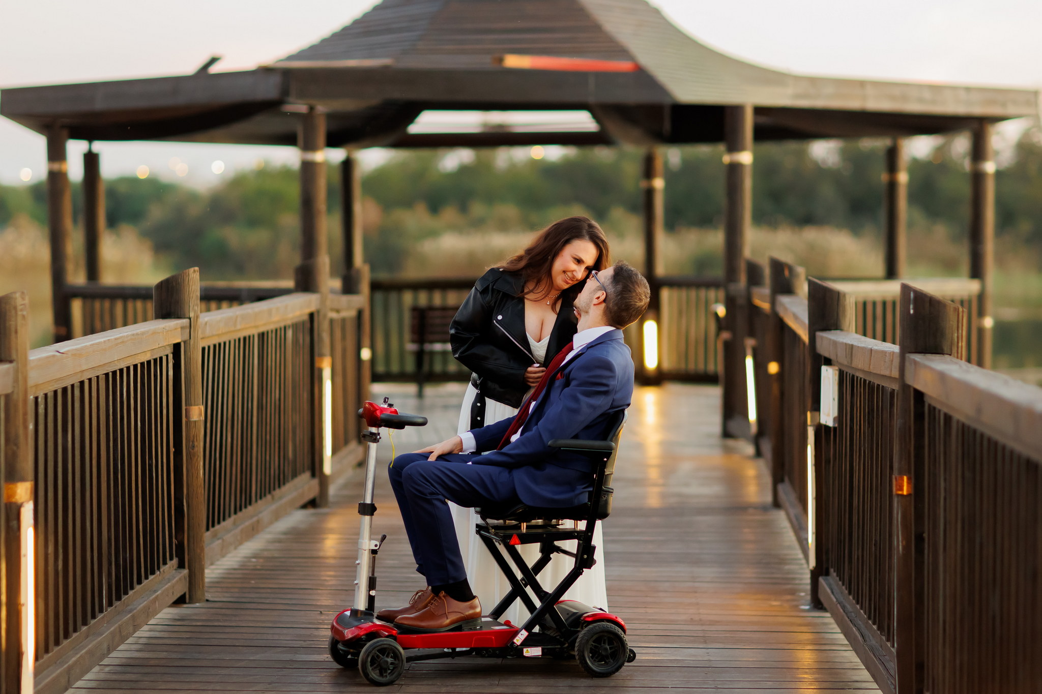 A romantic couple shares a tender moment on a wooden boardwalk at sunset. The woman, with long brown hair, wearing a black leather jacket over a white dress, leans intimately towards the man. The man, in a blue suit, white shirt, and red tie, sits comfortably in a red and silver mobility scooter, looking up at her with a warm smile. They are positioned centrally on the wooden structure, with a large, ornate wooden gazebo behind them and soft green foliage and a body of water in the blurred background, all bathed in the warm, golden light of the setting sun.
