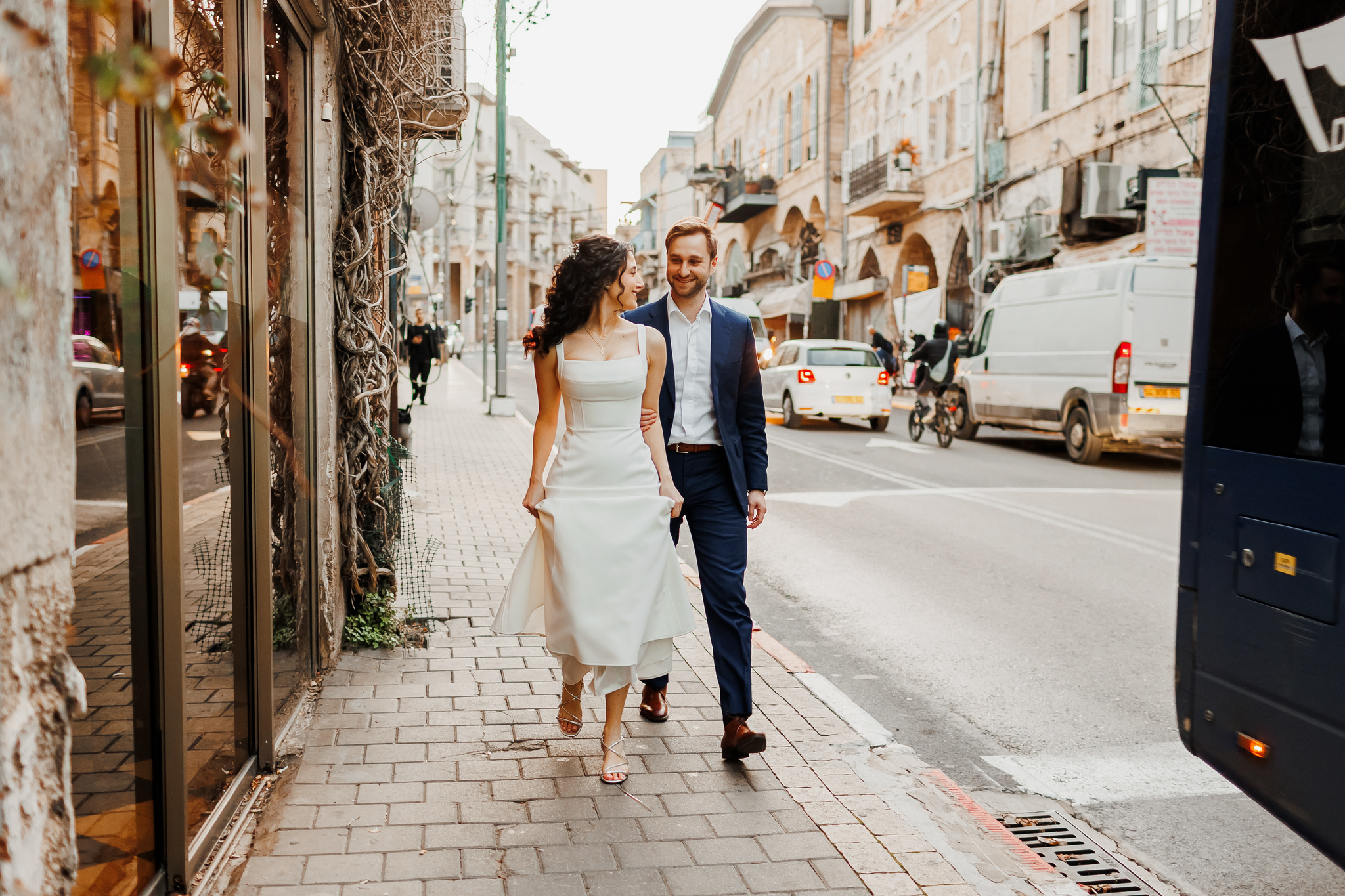 A smiling couple, dressed in a white wedding-style dress and a navy suit, walks arm-in-arm down a sunlit, cobblestone city sidewalk. The woman, with dark curly hair, looks up at the man, who is also smiling. To their left are reflective shop windows covered in intertwined vines. To their right and in the background, a busy street features old, ornate stone buildings, various vehicles including a white van and a scooter, and a partial view of a dark blue bus.
