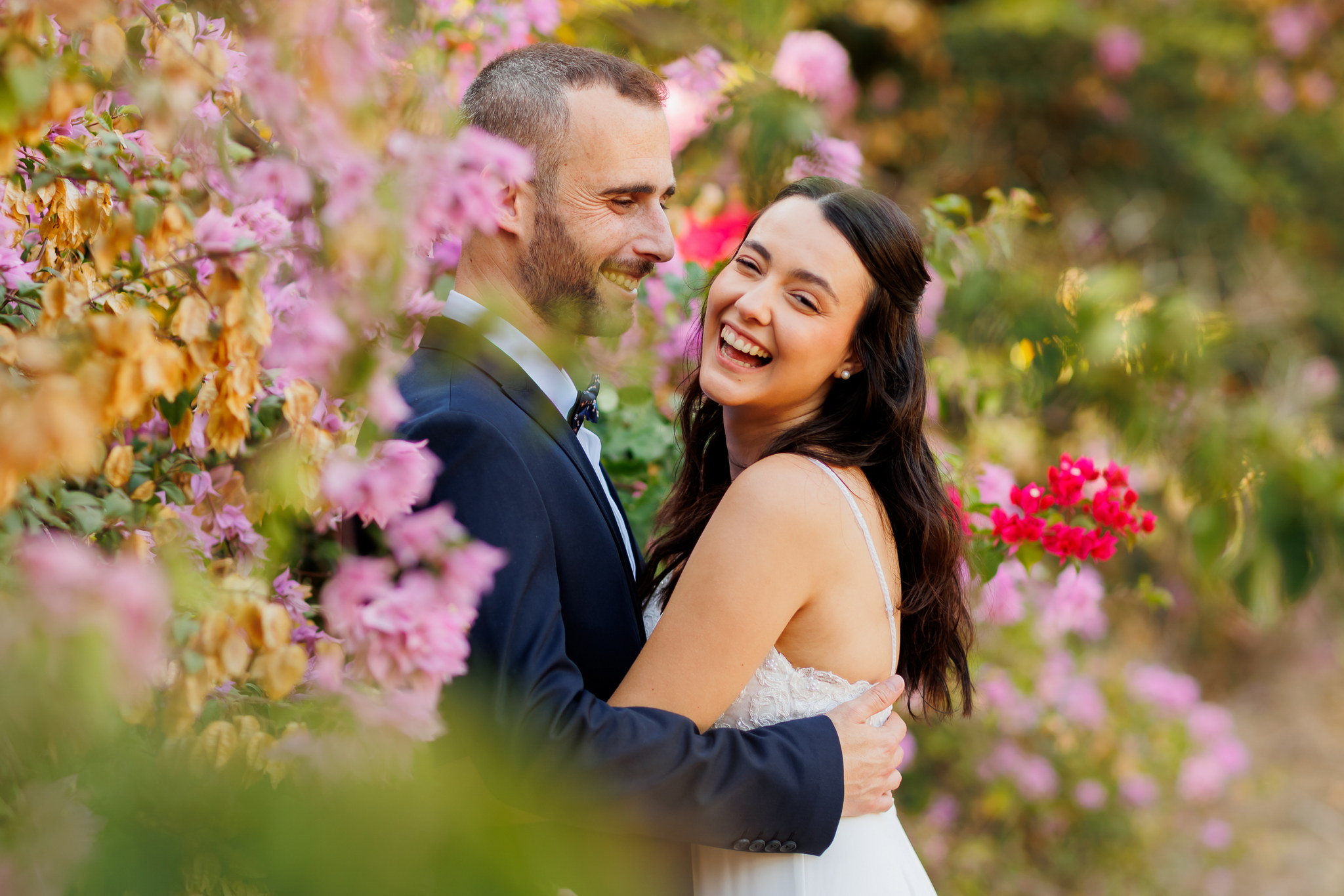 A joyful bride and groom embracing and laughing in a lush garden filled with vibrant pink and red bougainvillea flowers. The groom, wearing a dark blue suit and bow tie, looks happily at his bride, who is in a white, strappy wedding gown with a lace bodice, and has long dark hair and pearl earrings. The background is a soft, blurred tapestry of colorful flowers and green leaves, creating a natural and romantic atmosphere.