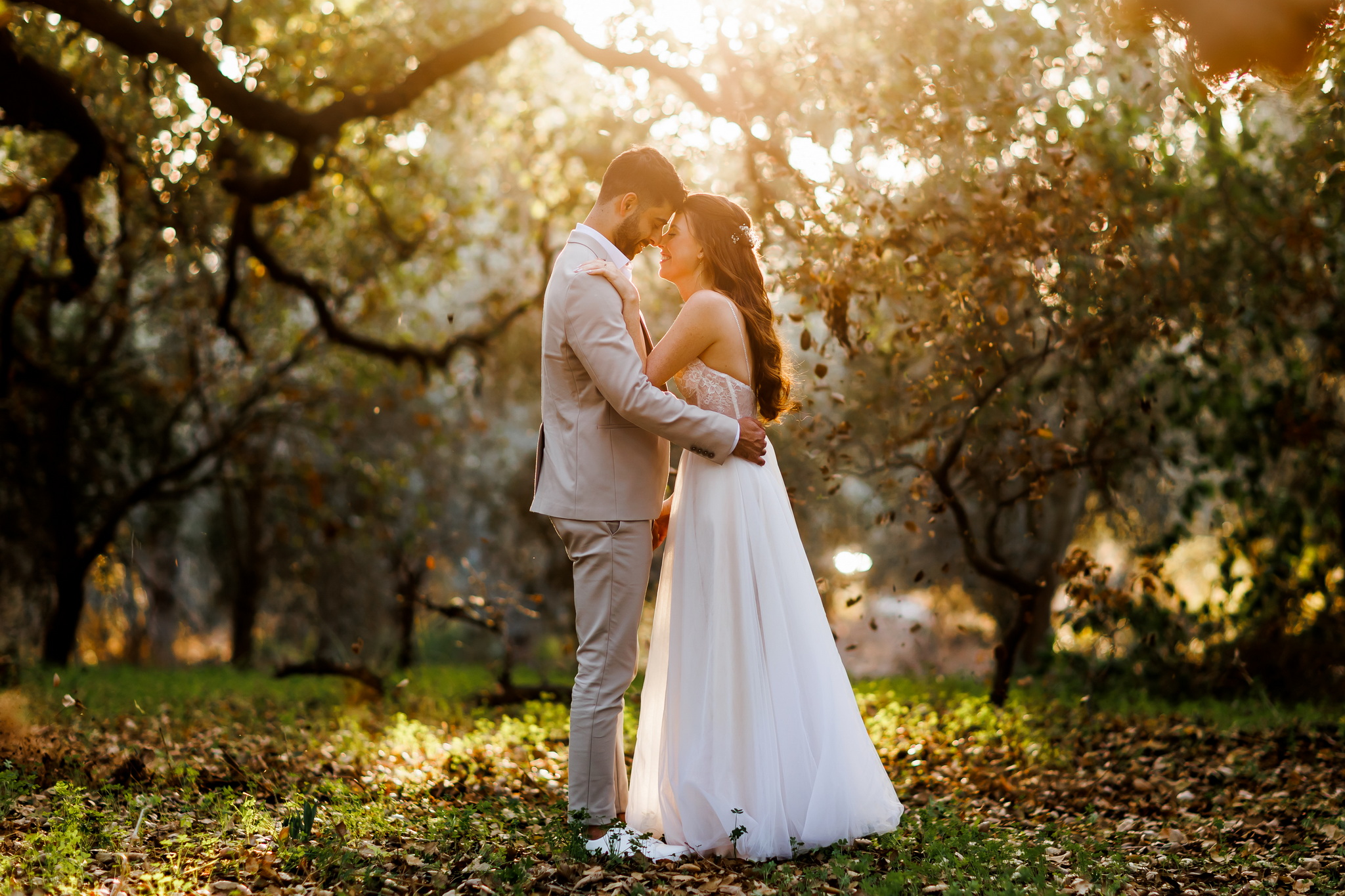 A romantic wedding couple embraces intimately in a sun-drenched autumnal park. The groom, in a light beige suit and white shirt, gently holds the bride, who wears a flowing white strapless gown with a delicate lace bodice. Their heads are touching, eyes closed, and soft smiles on their faces convey a moment of tender affection and joy. Golden sunlight streams through the canopy of trees behind them, creating a warm, ethereal glow and a beautiful bokeh effect. The ground is covered with fallen brown leaves and patches of green grass, bathed in the golden light, with some leaves seemingly suspended in the air around them.