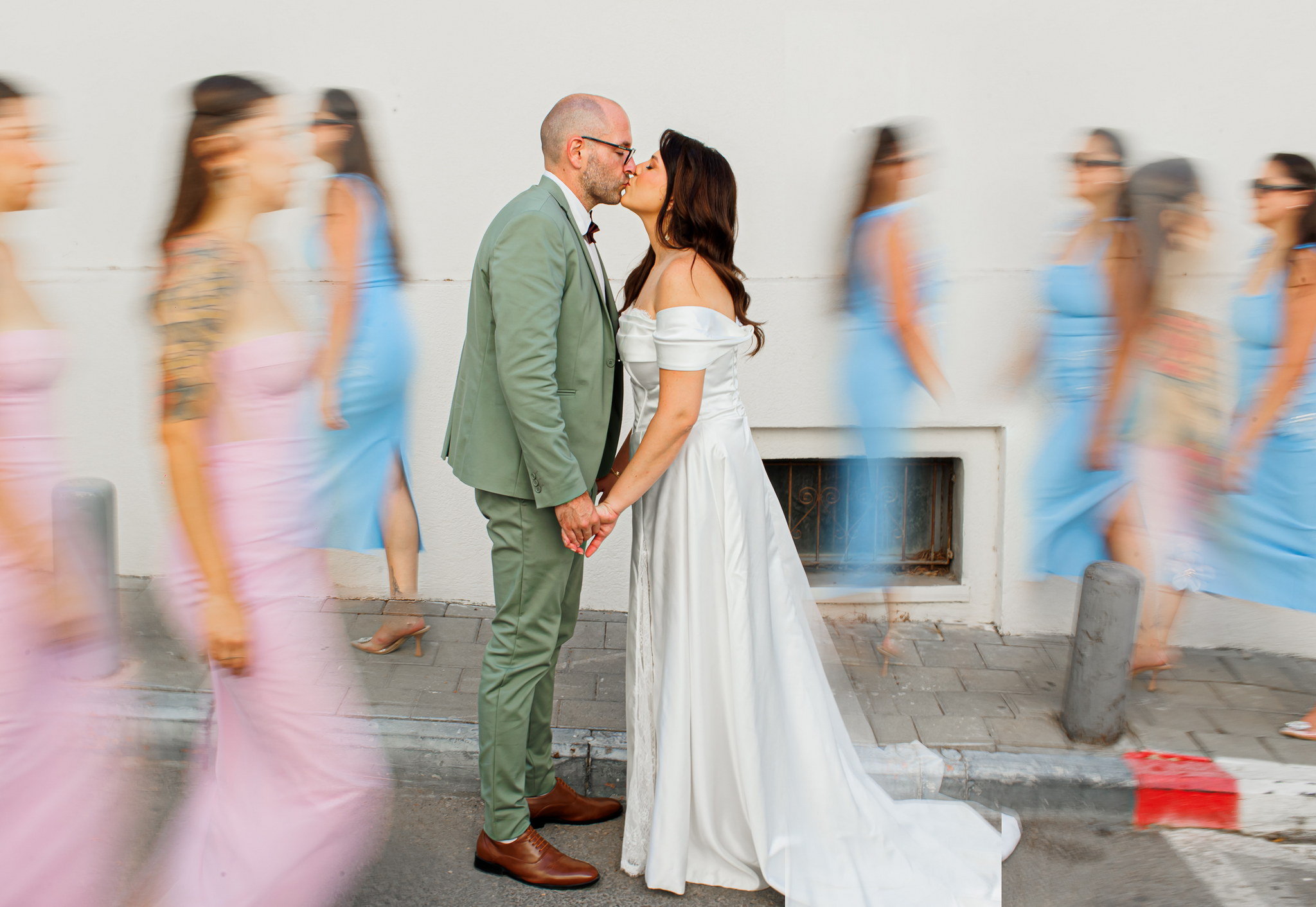 A newlywed couple shares a kiss on a paved sidewalk, holding hands, against a plain white building wall. The groom, a bald man with glasses, wears a light green suit with a dark red bowtie. The bride, with long dark hair, is in an elegant off-the-shoulder white wedding gown with a flowing train. On both sides of the couple, motion-blurred figures in pink and blue dresses create a dynamic background, suggesting a bustling scene around their intimate moment.